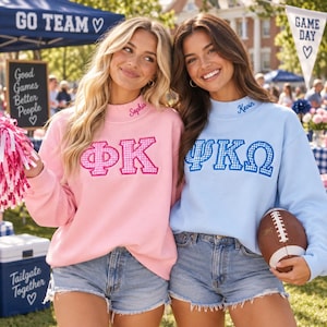 May include: Two long-sleeve crewneck sweatshirts, one light pink with pink gingham Greek letters and the name Sophia, and one light blue with blue gingham Greek letters and the name Kevin, both paired with light wash denim shorts.