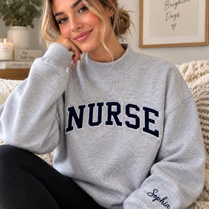 Peut inclure: Un sweat-shirt gris clair à col montant avec le mot NURSE inscrit en lettres capitales bleu marine sur la poitrine. Le poignet de la manche gauche est brodé du prénom Sophia en écriture cursive bleu marine. Le vêtement possède un col montant.