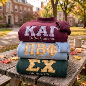 Könnte beinhalten: Ein Stapel aus drei Sweatshirts mit Rundhalsausschnitt. Das oberste ist kastanienbraun mit weiß-blau karierten griechischen Buchstaben KΔΓ und dem Text Delta Gamma. Das mittlere ist hellblau mit gelb karierten Buchstaben ΠΒΦ und dem Text Pi Beta Phi. Das unterste ist dunkelgrün mit gelb karierten Buchstaben ΣΧ und dem Text Sigma Chi.