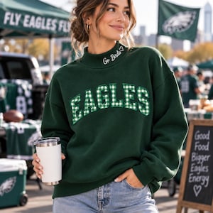 Könnte beinhalten: Ein dunkelgrünes Langarm-Sweatshirt mit Stehkragen, der mit dem Text Go Birds und einem Herz bestickt ist. Auf der Brust befindet sich das Wort EAGLES in großen, bogenförmigen Applikationen aus grün-weiß kariertem Stoff.