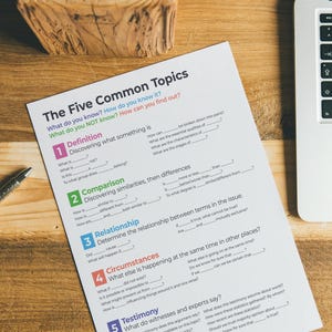 May include: A worksheet titled "The Five Common Topics" is printed on paper and placed on a wooden surface. The worksheet features numbered sections with fill-in-the-blank questions. A laptop and pen are also present.