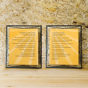 May include: Two framed prints with text from the Bible. The text is in English and Latin, set against a yellow background. The frames are black and rectangular, and the prints are displayed on a wooden surface.