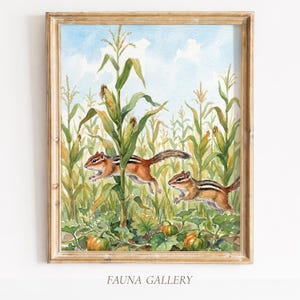 Puede incluir: Una pintura en acuarela enmarcada que representa a dos ardillas listadas de color marrón y blanco corriendo a través de un campo de maíz verde con tallos de maíz visibles y calabazas naranjas en el suelo bajo un cielo azul claro.