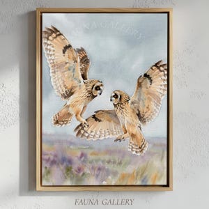 May include: Watercolor painting of two owls in flight, set against a soft blue and purple landscape. The owls have brown and white feathers, with wings spread wide. The artwork is framed in a natural wood frame, with "FAUNA GALLERY" printed at the bottom.