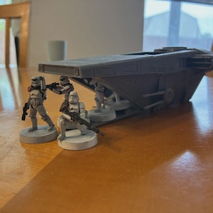 May include: A gray model of a transport vehicle with several Stormtrooper figures. The figures are on gray circular bases and are holding black rifles. The vehicle has a flat top and appears to be made of plastic. The figures are in various poses.
