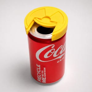 Puede incluir: Una lata roja de Coca-Cola con una tapa amarilla. La lata tiene el logotipo de Coca-Cola en blanco y las palabras "RECYCLE ME SEE YOU AGAIN SOON!" impresas en el lateral. La tapa amarilla est&aacute; dise&ntilde;ada para cubrir la abertura de la lata.
