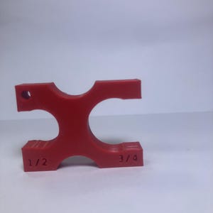 May include: A bright red, 3D-printed tool with a unique shape. It features a central opening and two curved sections, with the markings "1/2" and "3/4" on the sides. The tool has a small hole on one end.