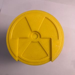 Puede incluir: Un objeto circular amarillo brillante con un s&iacute;mbolo de radiaci&oacute;n. El s&iacute;mbolo se compone de tres aspas que irradian desde un punto central, con un peque&ntilde;o c&iacute;rculo en el centro. El objeto tiene una superficie texturizada.