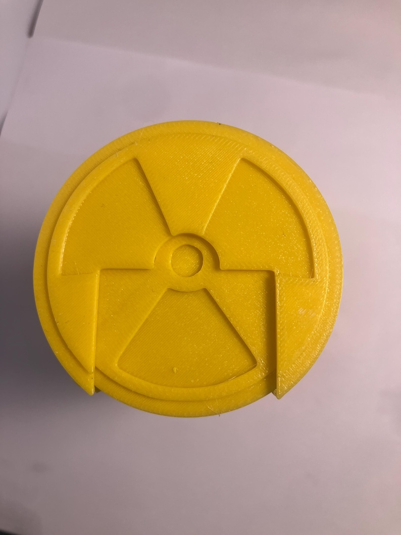 Puede incluir: Un objeto circular amarillo brillante con un s&iacute;mbolo de radiaci&oacute;n. El s&iacute;mbolo se compone de tres aspas que irradian desde un punto central, con un peque&ntilde;o c&iacute;rculo en el centro. El objeto tiene una superficie texturizada.