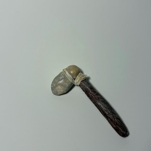 May include: A miniature stone axe with a dark brown wooden handle. The gray stone head is secured to the handle with a beige twine. The axe is a replica of a prehistoric tool.