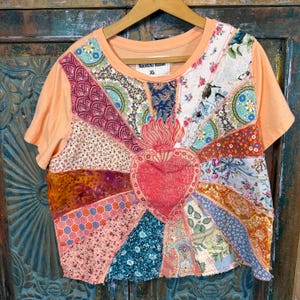 Op de afbeelding: Perzikkleurig T-shirt met een patchwork ontwerp. Het patchwork heeft verschillende bloemen- en patronenstoffen gerangschikt in een zonnestraalpatroon rond een rood hart met een vlam. Het shirt heeft korte mouwen en een ronde hals. Het label luidt "NOUVEAU REDUX XL".