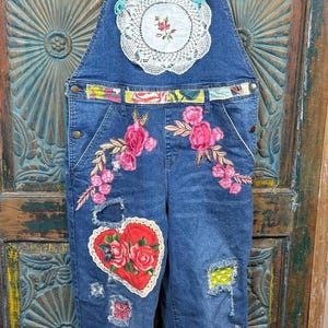 Könnte beinhalten: Blaue Jeans-Latzhose mit Blumenstickerei, Aufnähern und einer Herzapplikation. Die Latzhose hat ein Spitzendeckchen-Detail und bunte Träger. Ein einzigartiges, handgefertigtes Kleidungsstück.