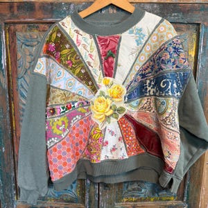 May include: A unique olive green sweatshirt with a patchwork design. The front features a sunburst pattern of various fabric scraps, including floral and geometric prints. A large appliqué of yellow roses is centered on the design. The sleeves and neckline are solid olive green.