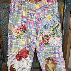May include: Pants featuring a multi-colored plaid pattern in pastel shades of pink, blue, yellow, and green. The garment is decorated with various floral appliques, white lace doilies, and a graphic print of a brown kitten with a pink bow.
