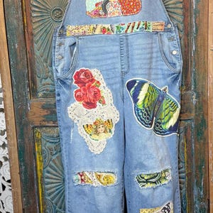 May include: Light blue denim overalls with colorful patchwork and decorative appliques. Features include a butterfly, roses, and a vintage-style image. The overalls have a relaxed fit and are embellished with floral and patterned fabrics.