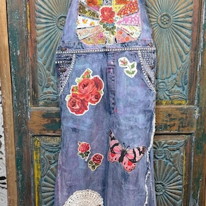 May include: Denim overalls with colorful floral and butterfly patches. The blue overalls feature a bib pocket with a patchwork design and decorative embellishments. The straps and edges are trimmed with patterned fabric, and the overall design is bohemian.