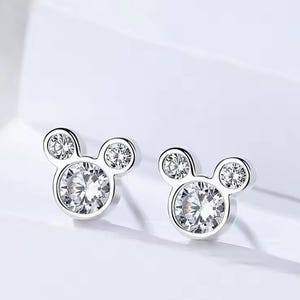 May include: A pair of silver-toned stud earrings shaped like a mouse head silhouette. Each earring features one large round clear faceted stone in the centre and two smaller round clear faceted stones forming the ears, all set in a polished metal frame.