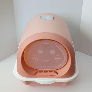 May include: A pink and white cat litter box with a rounded top and a cat-face-shaped entry door. The litter box has a light pink body, a darker pink accent, and a white base. The entry door has a clear plastic cover.