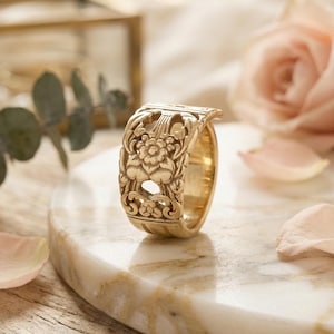 Puede incluir: Un anillo dorado con un intrincado diseño floral. El anillo se exhibe sobre una superficie de mármol, con un fondo borroso que presenta una rosa y otros elementos decorativos. Las detalladas tallas del anillo son el punto focal.