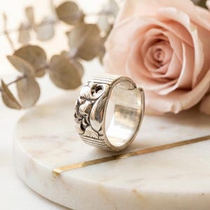 Può includere: Un anello in argento con una fascia larga dalla finitura lucida, caratterizzato da motivi floreali o organici in rilievo al centro. I bordi superiore e inferiore della fascia sono decorati con sottili linee verticali parallele.