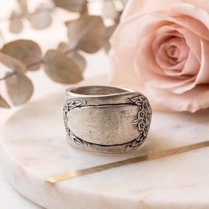 May include: A silver-toned metal ring crafted from a repurposed spoon handle. The band features a wide, smooth central panel flanked by intricate embossed floral and leaf patterns on both sides, showcasing a weathered, antique-style finish.