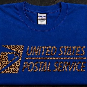 Könnte beinhalten: Ein königsblaues T-Shirt mit Rundhalsausschnitt, das das Logo und den Schriftzug United States Postal Service in einem braun-schwarzen Leopardenmuster zeigt. Das Gildan-Markenetikett ist am inneren Halsausschnitt sichtbar.
