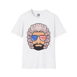 May include: White t-shirt featuring a cartoon illustration of a man with a powdered wig, sunglasses with the American flag design, and a black beard. The design is centered on the front of the shirt.
