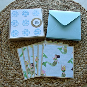 May include: A set of six blank greeting cards with a mermaid theme. The cards are decorated with mermaids with green tails and colorful tops. The cards are packaged with a set of blue envelopes and a sheet of stickers with a blue and white wave pattern.