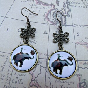 May include: A pair of bronze earrings with a knotted design and a round pendant featuring a white background with a black and white illustration of an elephant with a person riding on top.