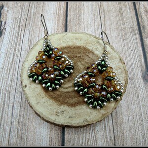 May include: A pair of handcrafted beaded earrings. Each earring features a diamond-shaped design with amber and green beads, accented with silver-colored beads. The earrings are displayed on a wooden slice.