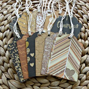 May include: A set of 10 gift tags with a variety of patterns and textures. The tags are made of brown cardstock and feature gold glitter, stripes, and hearts. The tags are tied with twine and have a rustic, vintage look.