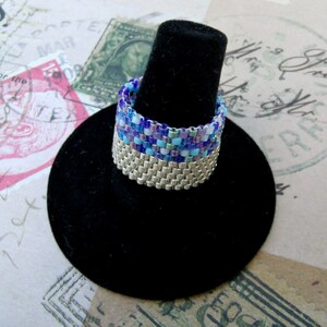 May include: A silver and blue beaded ring on a black ring display stand. The ring is made of small, square beads in shades of blue, purple, and white.