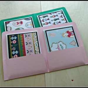 May include: A pink paper folder with green and pink pockets, containing four Christmas cards. The cards display festive designs such as nutcrackers, baubles, and candy canes. The cards have black borders.