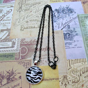 May include: A black chain necklace with a round pendant featuring a zebra print design. The pendant has a silver-colored bezel. The necklace is displayed on a background of vintage paper ephemera.