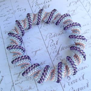 Puede incluir: Un brazalete de cuentas en espiral con cuentas moradas, blancas y rosadas. El brazalete está hecho de pequeñas cuentas redondas que están enhebradas en un patrón en espiral. Las cuentas son iridiscentes y tienen un brillo sutil.