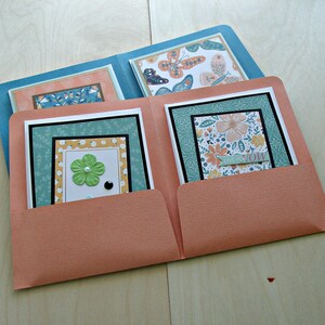 Puede incluir: Una carpeta de color melocotón con dos bolsillos contiene cuatro tarjetas con diseños florales y de mariposas. Las tarjetas tienen un esquema de color turquesa, verde y blanco. El texto en una tarjeta dice "Live in the Now".