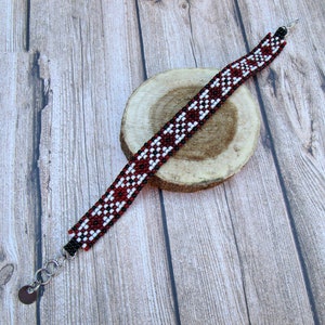 May include: A red, white, and black beaded bracelet with a geometric pattern. The bracelet is on a wooden surface.