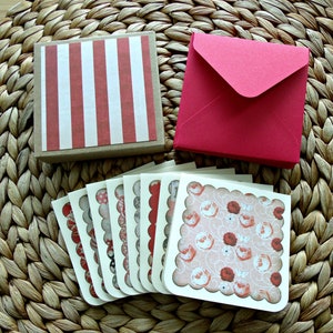 May include: A set of six blank greeting cards with scalloped edges and a floral pattern in shades of pink and red. The cards are packaged in a brown box with red and white stripes and come with a red envelope.