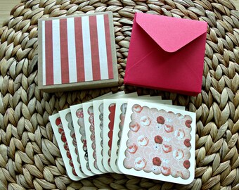 Pode incluir: Um conjunto de seis cartões de felicitação em branco com bordas festoneadas e um padrão floral em tons de rosa e vermelho. Os cartões são embalados numa caixa castanha com riscas vermelhas e brancas e vêm com um envelope vermelho.
