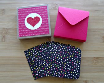 Op de afbeelding: Een verzameling briefpapier op een houten oppervlak. Bevat een kleine, vierkante doos met een roze en wit patroon en een rood glitterhart. Er zijn ook verschillende zwarte kaarten met kleurrijke driehoekpatronen en een stapel roze enveloppen.