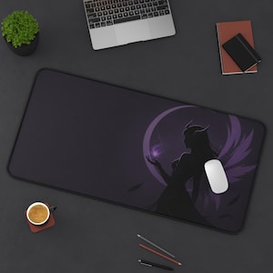 May include: A large, rectangular black mousepad with a purple and black graphic of a silhouetted figure with wings and a crescent moon. A white mouse sits on the pad. A laptop, coffee cup, and stationery are also visible.