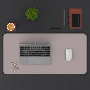 May include: A desk mat with a floral design, a laptop, and a mouse. The mat is a muted pink color. A cup of coffee, a small plant, a notebook, and a pen are also on the desk.