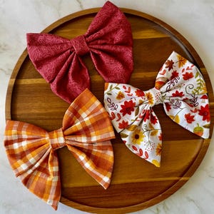 May include: Three fabric hair bows arranged on a wooden tray. One bow is solid dark red with a paisley pattern, one is orange and white plaid, and the third is white with a colourful print of autumn leaves, flowers, and small mushrooms.