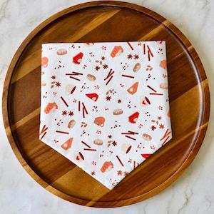 May include: A white triangular fabric bandana featuring a repeating pattern of orange and red autumn-themed illustrations, including slices of pumpkin pie, cinnamon sticks, star anise, coffee mugs, and small round pastries scattered across the surface.