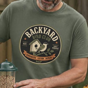 Puede incluir: Un hombre con una camiseta verde con un diseño de "Backyard Bird Club" llena un comedero para pájaros verde con semillas. Un nido y unos prismáticos son visibles en el fondo. El comedero es cilíndrico.