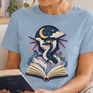 Può includere: Maglietta azzurra con un disegno di drago, una luna crescente, stelle e un libro aperto. Il drago è blu scuro con ali viola. Una persona indossa la maglietta e legge un libro. Una tazza bianca con la scritta "Just One More Chapter" è visibile.