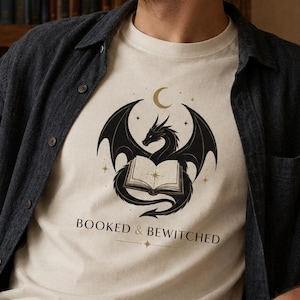 Può includere: T-shirt color crema con un disegno di drago nero, una luna crescente e stelle sopra un libro aperto. Il testo "BOOKED & BEWITCHED" è sotto. La maglietta è indossata con una camicia blu scuro abbottonata.