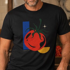 Peut inclure: Un t-shirt noir à manches courtes avec une impression graphique représentant une tomate rouge à tige verte, un rectangle vertical bleu, un demi-cercle jaune et une petite étoile blanche à quatre branches. Le texte TOMATO N° 5 est imprimé.
