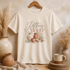 Può includere: T-shirt color crema con grafica a tema ceramica. Il design include vari vasi, ciotole e un piatto in tonalità marroni e rosa, con le parole "Pottery Mood" scritte in elegante corsivo.