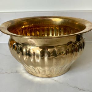 May include: A polished brass bowl featuring a wide, flared rim and a rounded body with vertical fluted indentations. The metallic surface has a warm, golden tone with subtle variations in luster and texture across the exterior and interior.
